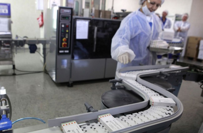 Operaria con cofia y bata estéril supervisa cajas de medicamento en una banda transportadora