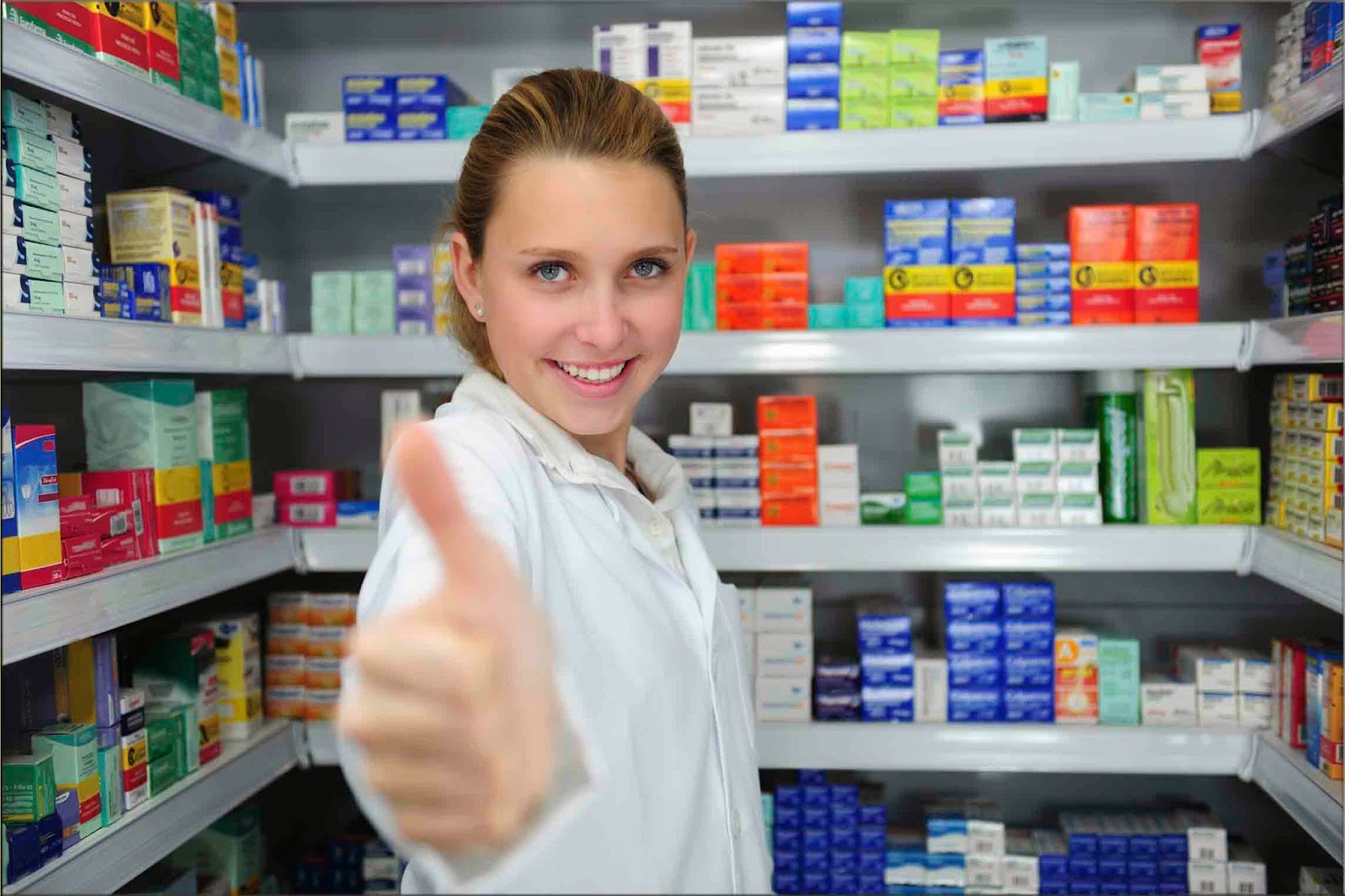 Mujer con bata blanca levanta el pulgar frente a anaqueles llenos de cajas de medicamentos
