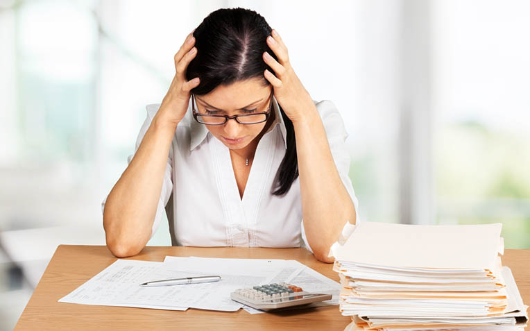 Mujer con las manos en la cabeza ante papeles, una calculadora y una pila de expedientes