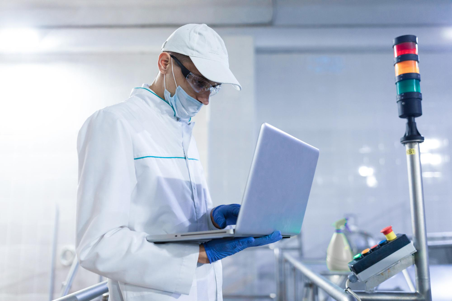 Operario con gorra, cubrebocas, lentes y guantes usa una computadora portátil en un área de producción