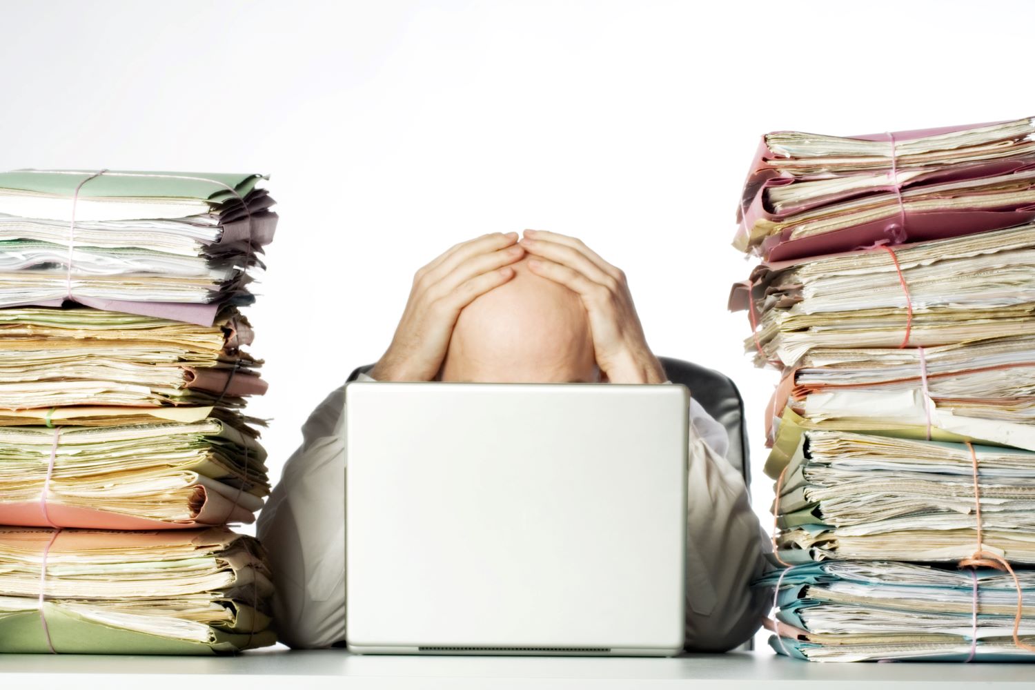 Hombre con la cabeza entre las manos frente a una laptop, entre dos pilas altas de expedientes